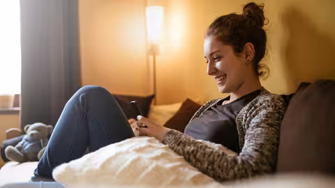 Foto: Eine junge Frau, die ihre Haare zu einem Dutt zusammengebunden hat, sitzt auf der Couch und schaut lächelnd auf das Display ihres Smartphones.