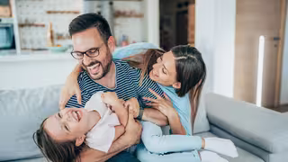 Foto: Zwei junge Eltern albern mit ihrer Tochter auf der Couch herum.