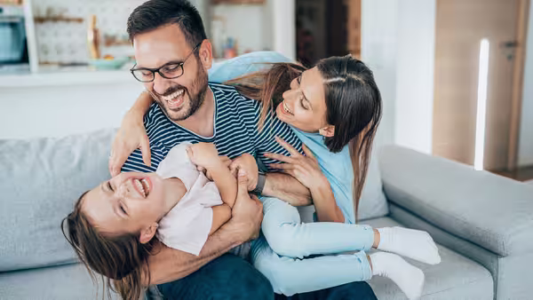 Foto: Zwei junge Eltern albern mit ihrer Tochter auf der Couch herum.