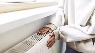 Eine Person legt beide Hände auf einen warmen Heizkörper unter einem Fenster. Sie trägt einen hellen Pullover.