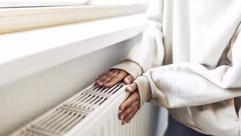 Eine Person legt beide Hände auf einen warmen Heizkörper unter einem Fenster. Sie trägt einen hellen Pullover.