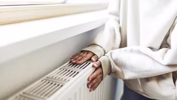 Eine Person legt beide Hände auf einen warmen Heizkörper unter einem Fenster. Sie trägt einen hellen Pullover.