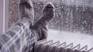 Person mit warmen Socken legt die Füße auf einen Heizkörper, im Hintergrund ein verregnetes Fenster.