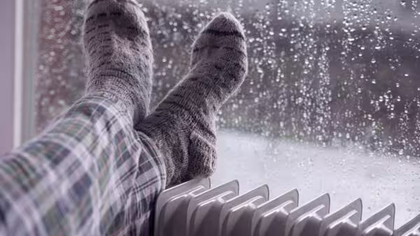 Person mit warmen Socken legt die Füße auf einen Heizkörper, im Hintergrund ein verregnetes Fenster.