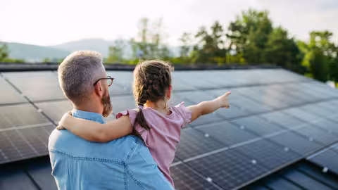 Foto: Rückansicht eines Vaters mit Vollbart, der seine kleine Tochter in rosafarbenem Kleid auf dem Arm hat, mit der zusammen er eine Photovoltaikanlage auf einem Dach betrachtet