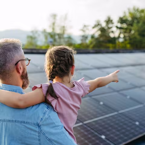 Foto: Rückansicht eines Vaters mit Vollbart, der seine kleine Tochter in rosafarbenem Kleid auf dem Arm hat, mit der zusammen er eine Photovoltaikanlage auf einem Dach betrachtet