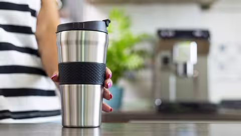 Foto: Die Hand einer jungen Frau im blau-weißgestreiften T-Shirt stellt einen Thermobecher auf einen Tisch, im Hintergrund steht ein Kaffeevollautomat.