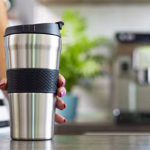 Foto: Die Hand einer jungen Frau im blau-weißgestreiften T-Shirt stellt einen Thermobecher auf einen Tisch, im Hintergrund steht ein Kaffeevollautomat.