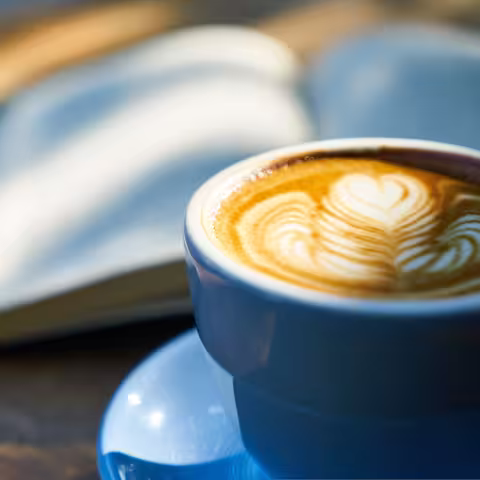 Foto: eine blaue Tasse Cappuccino vor einem aufgeschlagenen Buch auf einem Tisch