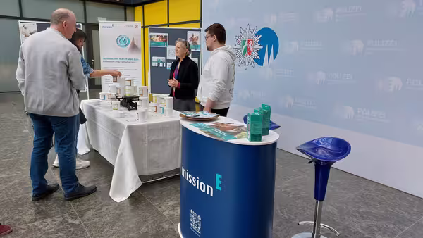 Zwei Personen besuchen einen Informationsstand und sind in Gesprächen eingebunden.