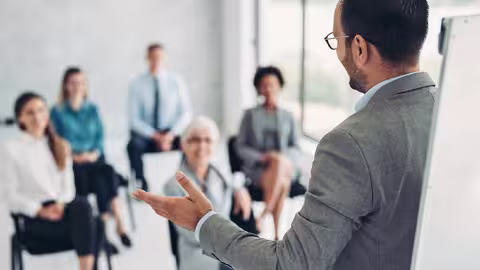 Foto: Ein Mann mit kurzen dunklen Haaren, Brille und hellgrauem Sakko spricht vor einem interessierten Publikum.