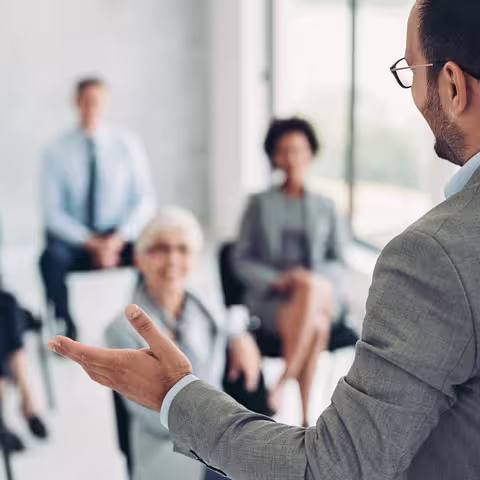 Foto: Ein Mann mit kurzen dunklen Haaren, Brille und hellgrauem Sakko spricht vor einem interessierten Publikum.