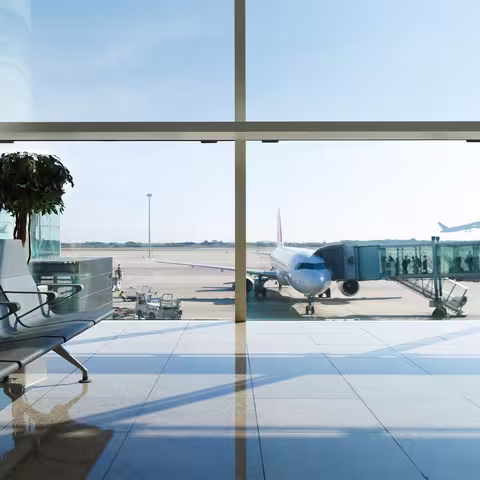 Foto: Blick aus einem leeren Sitzbereich eines Gates am Flughafen auf das Rollfeld mit einem Flugzeug in der Bildmitte