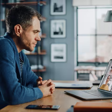 Foto: Ein junger blonder Mann im Jeanshemd sitzt vor seinem Notebook, auf dem andere Teilnehmende eines gemeinsamen Termins zu sehen sind.