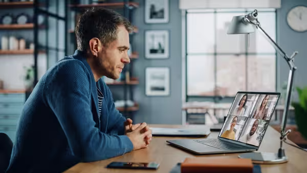 Foto: Ein junger blonder Mann im Jeanshemd sitzt vor seinem Notebook, auf dem andere Teilnehmende eines gemeinsamen Termins zu sehen sind.