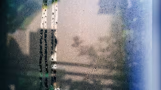 Beschlagenes Fenster mit vielen Wassertropfen und herunterlaufenden Spuren, durch das die Außenansicht nur verschwommen erkennbar ist.