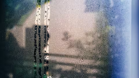 Beschlagenes Fenster mit vielen Wassertropfen und herunterlaufenden Spuren, durch das die Außenansicht nur verschwommen erkennbar ist.