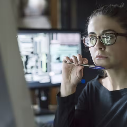Foto: Eine junge Frau mit Brille und Zopf sitzt vor ihrem großen PC-Bildschirm und hält einen Kugelschreiber ans Kinn.