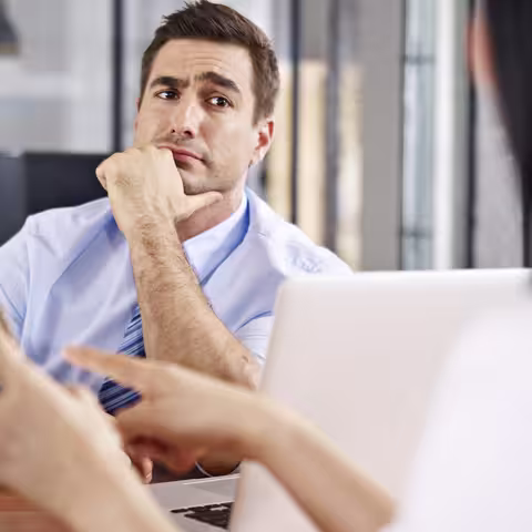 Foto: Ein Mann mittleren Alters sitzt mit skeptischer Miene vor einem Schreibtisch, hinter dem eine Frau sitzt, die ihm mithilfe eines Tablets etwas erläutert.
