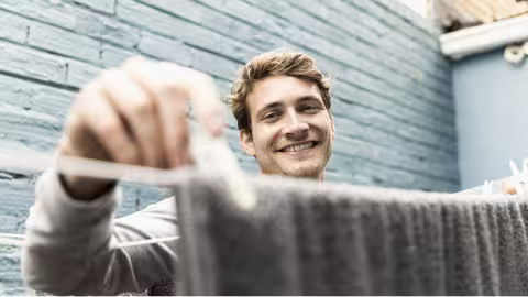 Foto: Ein junger, blonder lächelnder Mann trocknet Wäsche im Hinterhof.