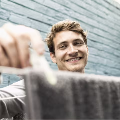 Foto: Ein junger, blonder lächelnder Mann trocknet Wäsche im Hinterhof.