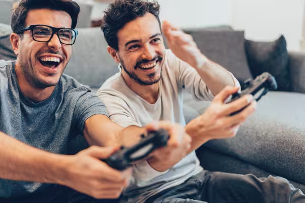 Zwei Personen sitzen auf einem Sofa und spielen gemeinsam Videospiele, beide lachen und halten Controller in den Händen.