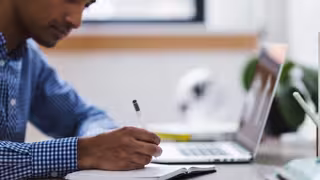 Mann sitzt am Schreibtisch vor seinem Laptop und schreibt mit einem Stift in ein Buch.