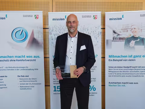 Foto: Thomas Hassel, ein schlanker Mann mit grauem Kinnbart, hält seine Auszeichnung in die Kamera.