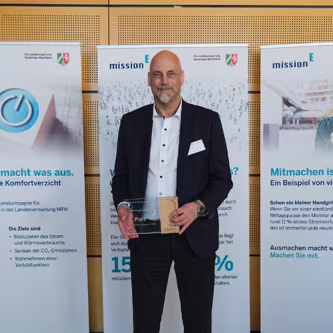 Foto: Thomas Hassel, ein schlanker Mann mit grauem Kinnbart, hält seine Auszeichnung in die Kamera.