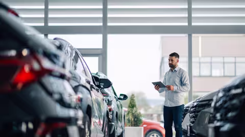 Foto: Ein junger Angestellter in hellgrauem Pullover und Jeans steht mit einem Klemmbrett vor einer Reihe neuer Autos im Verkaufsraum eines Händlers.