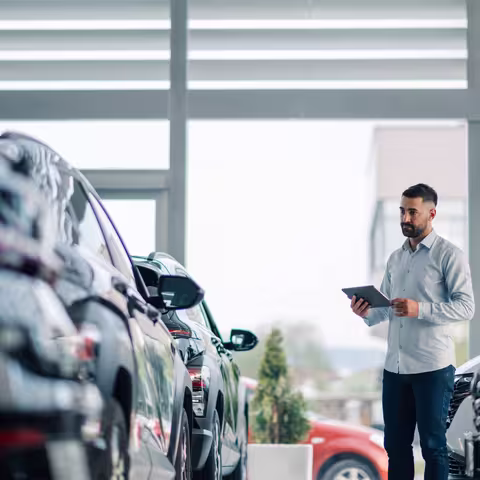 Foto: Ein junger Angestellter in hellgrauem Pullover und Jeans steht mit einem Klemmbrett vor einer Reihe neuer Autos im Verkaufsraum eines Händlers.