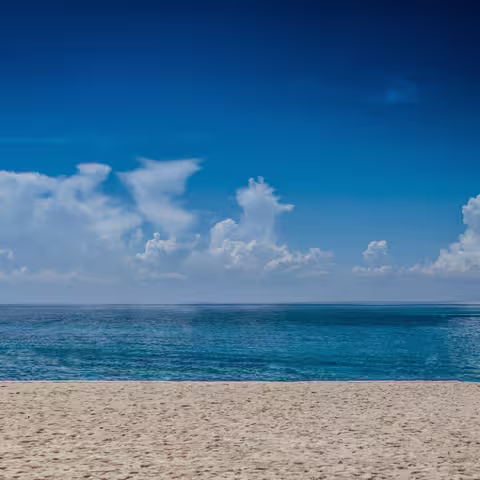 Foto: Blick von einem feinen, hellen Sandstrand auf das ruhige, hellblaue Meer, an dessen Horizont weiße Wolken stehen