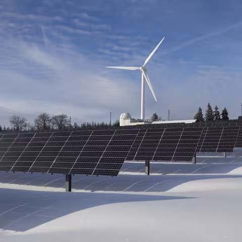 Foto: eine Freiflächensolaranlage vor einem Windrad in hügeliger winterlicher Schneelandschaft