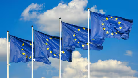 Foto: Vier Fahnen der Europäischen Union wehen vor blauem, leicht bewölktem Hintergrund.