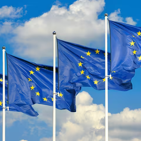 Foto: Vier Fahnen der Europäischen Union wehen vor blauem, leicht bewölktem Hintergrund.