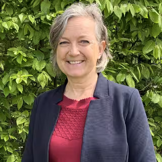 Foto: Frau Eichen steht vor einer hohen grünen Hecke, trägt schulterlanges graues Haar und einen dunklen Blazer über dem roten Shirt.