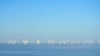 Mehrere weiße Windkraftanlagen stehen in einem ruhigen Offshore-Windpark auf dem Meer. Die Rotoren ragen aus dem Wasser und sind gleichmäßig in Reihen angeordnet. Der Himmel ist klar und blau, und die Wasseroberfläche wirkt glatt und leicht neblig, wodurch die Windräder besonders deutlich hervortreten.