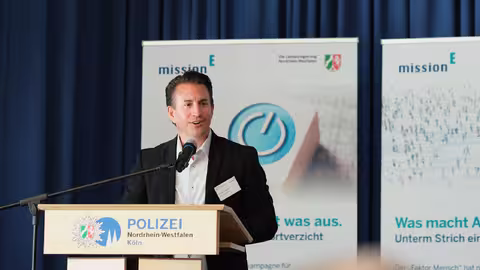 Foto: Christian Mildenberger, der Geschäftsführer von NRW.Energy4Climate, steht am Rednerpult mit dem Schriftzug des Polizeipräsidiums Köln. Hinter ihm ist ein dunkelblauer Vorhang zu erkennen.