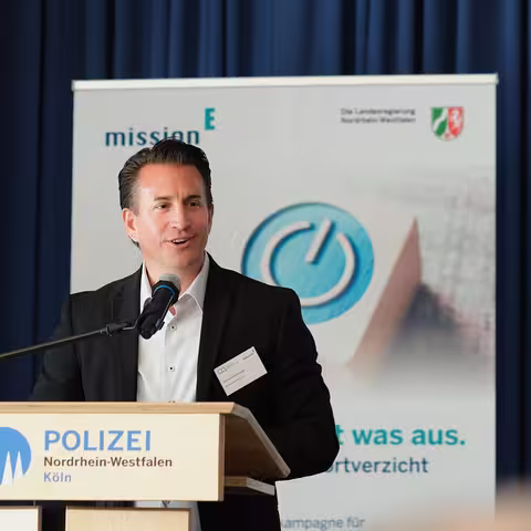 Foto: Christian Mildenberger, der Geschäftsführer von NRW.Energy4Climate, steht am Rednerpult mit dem Schriftzug des Polizeipräsidiums Köln. Hinter ihm ist ein dunkelblauer Vorhang zu erkennen.