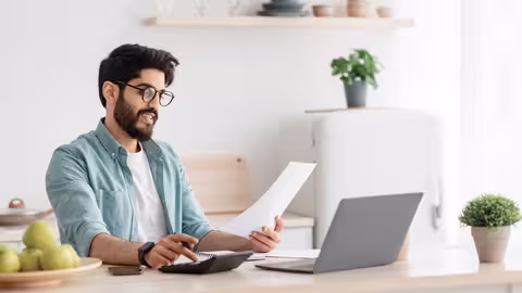 Foto: Ein junger, dunkelhaariger Mann mit Brille sitzt vor seinem Notebook und hält ein Blatt Papier in der Hand.
