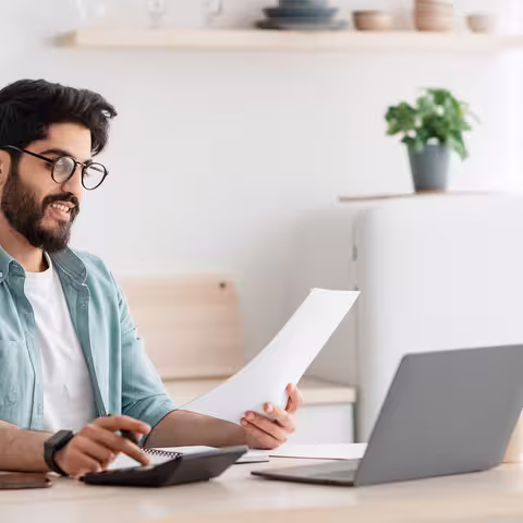 Foto: Ein junger, dunkelhaariger Mann mit Brille sitzt vor seinem Notebook und hält ein Blatt Papier in der Hand.