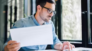 Person sitzt am Schreibtisch vor einem Laptop und hält ein Dokument, während sie konzentriert darauf schaut.