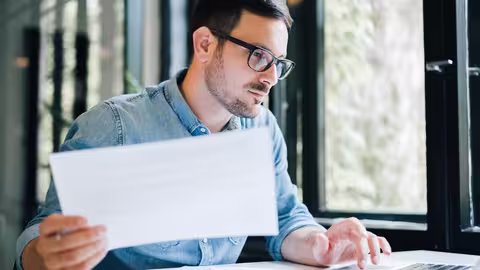Person sitzt am Schreibtisch vor einem Laptop und hält ein Dokument, während sie konzentriert darauf schaut.