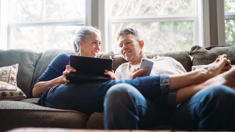 ein Paar mittleren Alters entspannt mit Tablet und Smartphone auf der Couch